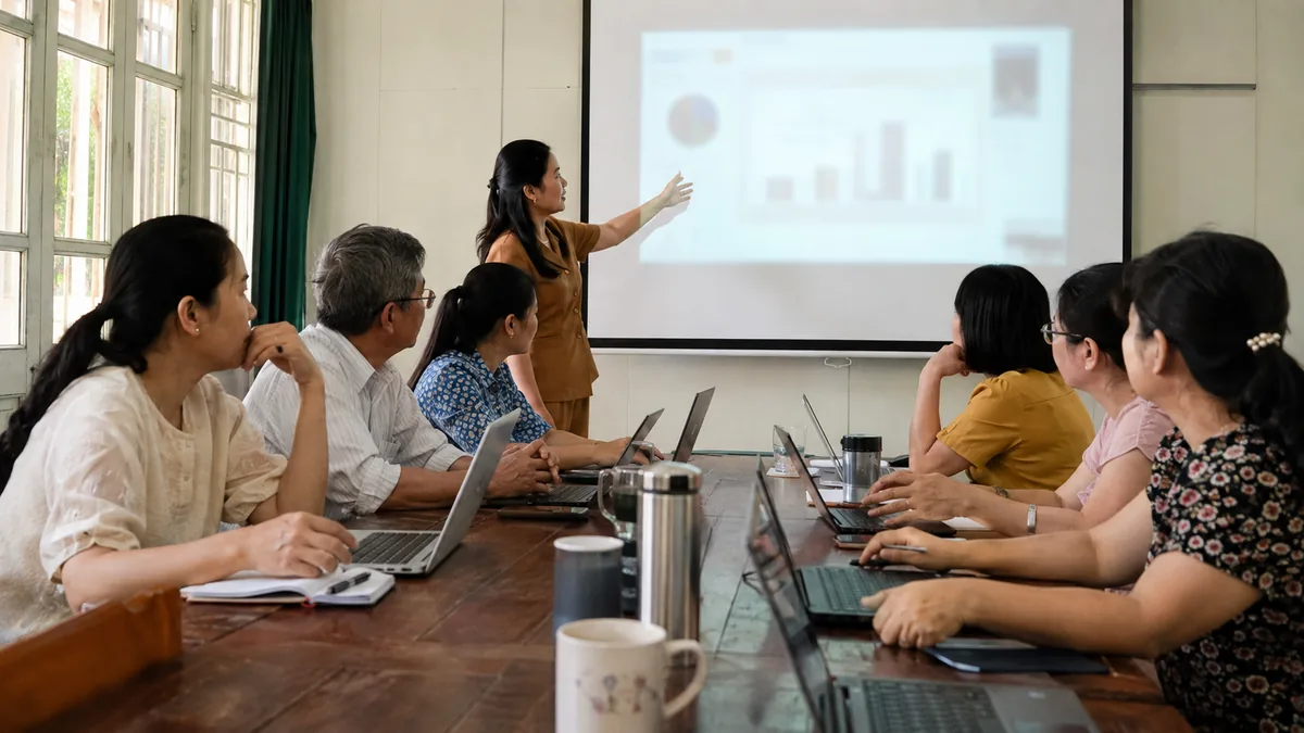 Buổi tập huấn giáo viên ứng dụng AI trong soạn giảng — một giáo viên trình bày trước màn chiếu, các đồng nghiệp ngồi quanh bàn với máy tính xách tay