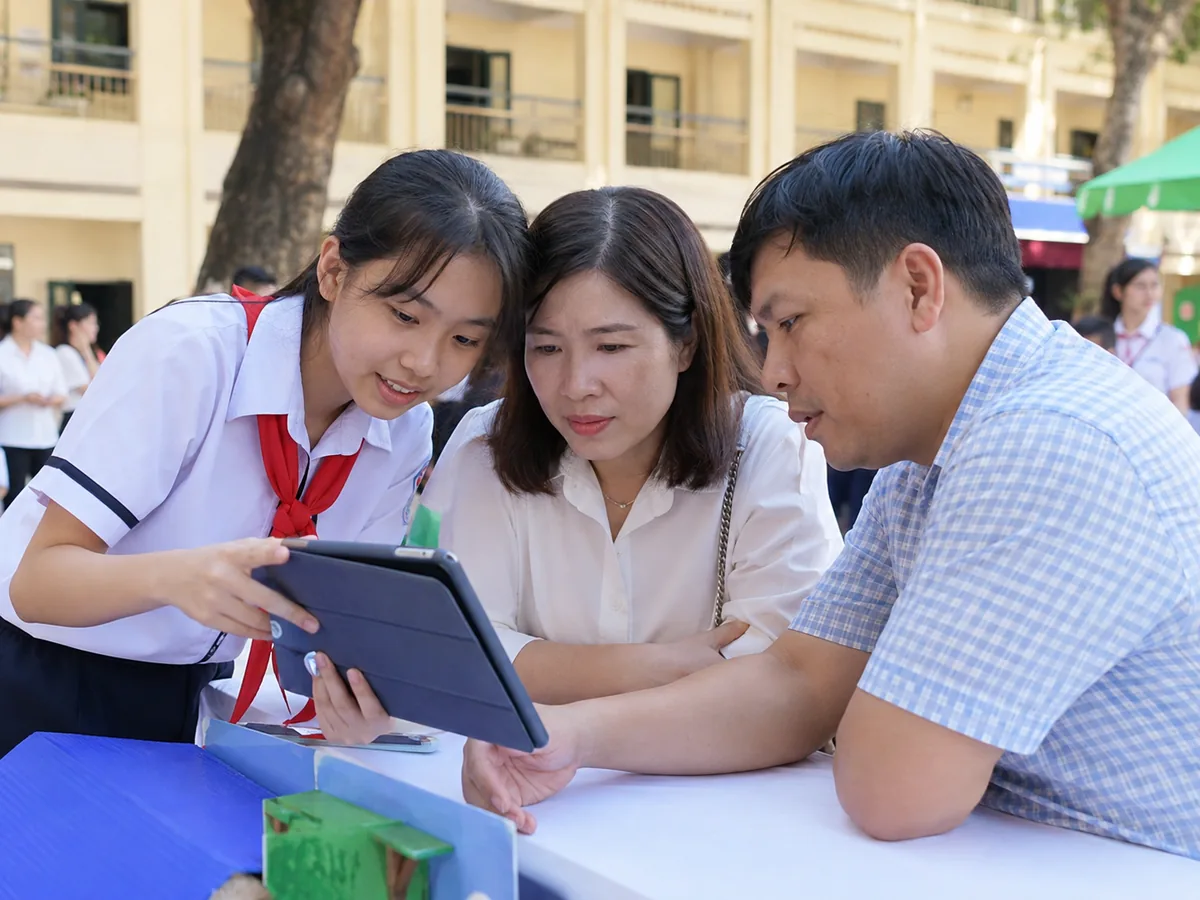 Một học sinh cầm máy tính bảng hướng dẫn cha mẹ cách dùng ứng dụng học tập trong sân trường